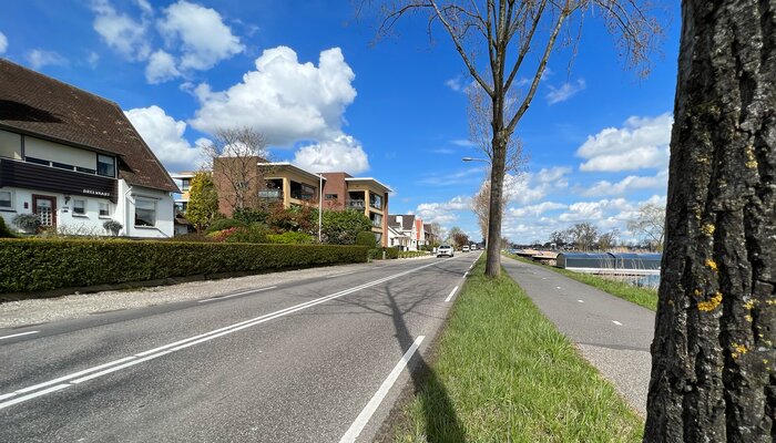 Een autoweg, met aan de linkerkant huizen en aan de rechterkant een apart fietspad. Naast het fietspad is het water van de Reeuwwijkse Plassen.
